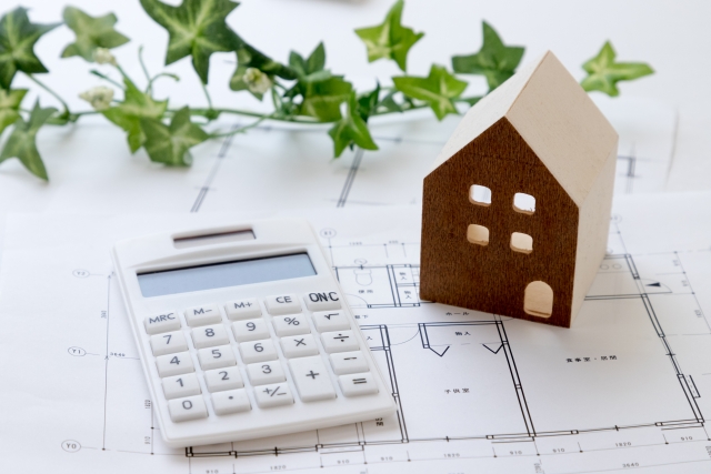 設計図の上に家の模型と電卓。住宅計画や費用計算を示すイメージ。